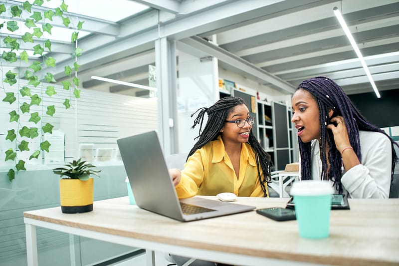 Two young women in office looking surprised at something they are seeing on laptop screen.